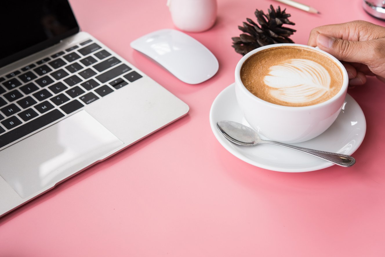 Hand holding a cup of coffee on the desk pink with a notebook computer.